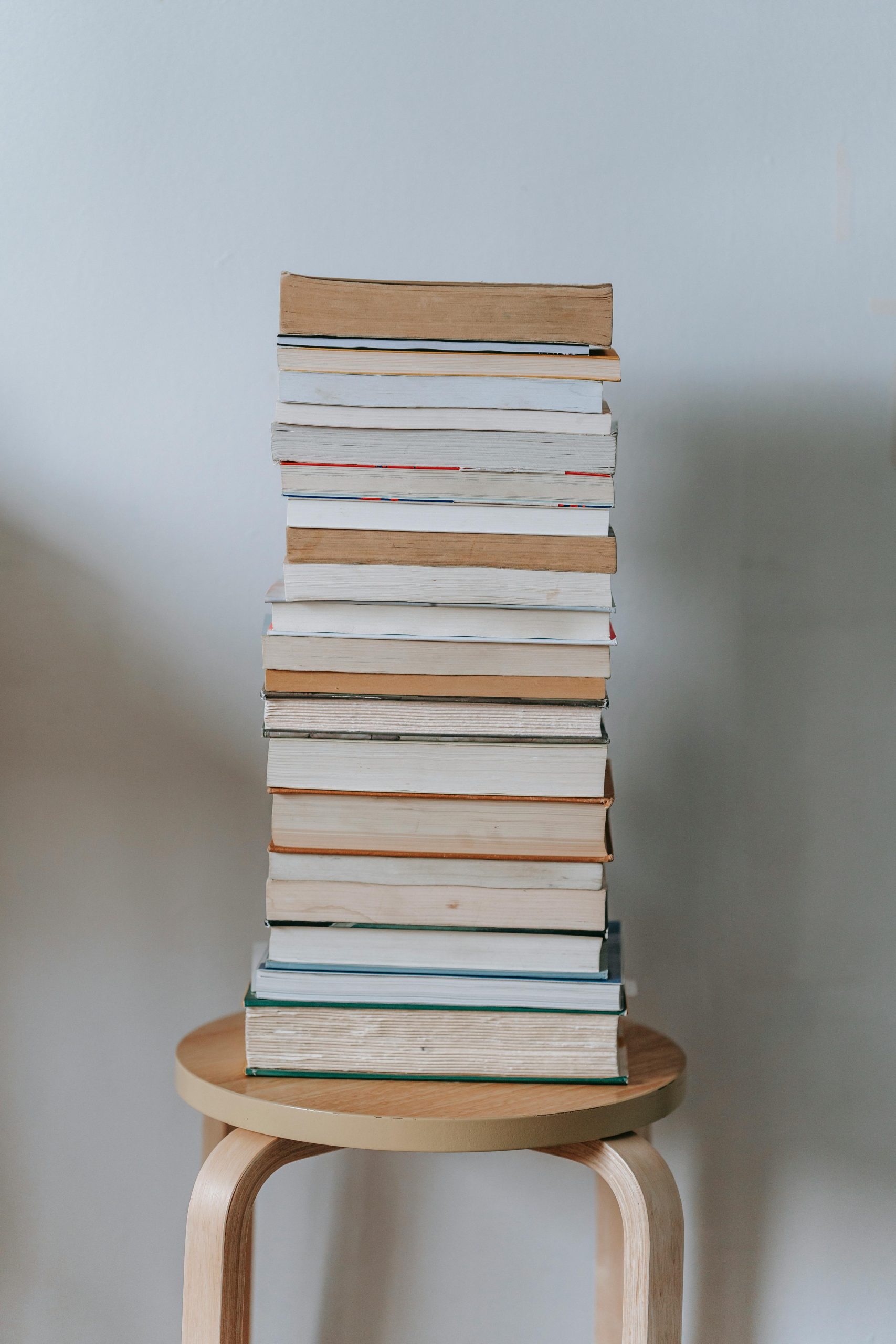 A neat pile of books stacked vertically on a wooden stool with a plain background.